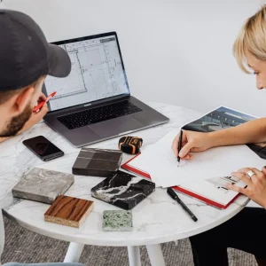 Two designers collaborating on a project, utilizing a laptop and drafting materials on a round table.